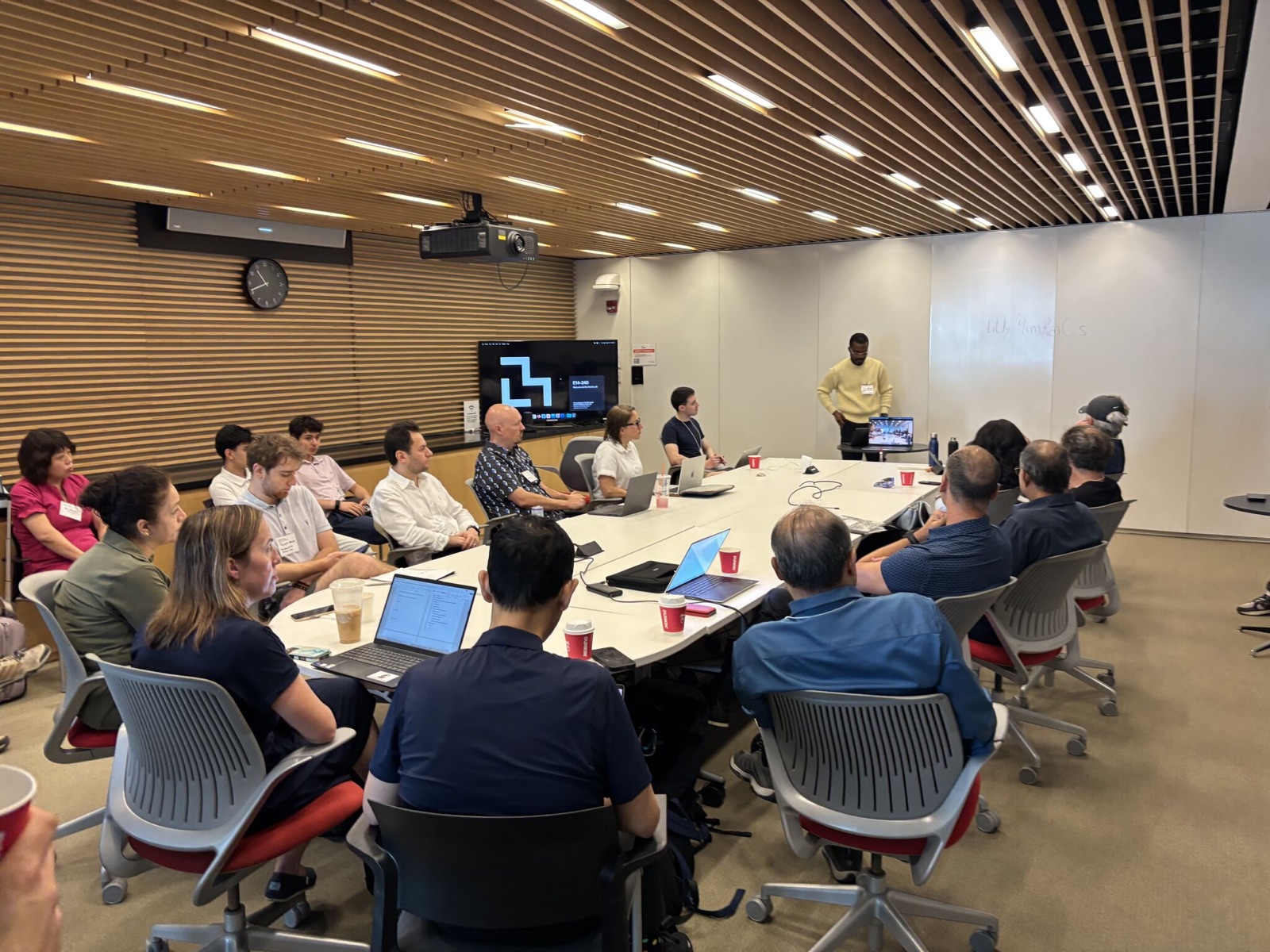Attendees seated around a large conference table with laptops, listening to a presenter at the front of a wood-panelled meeting room